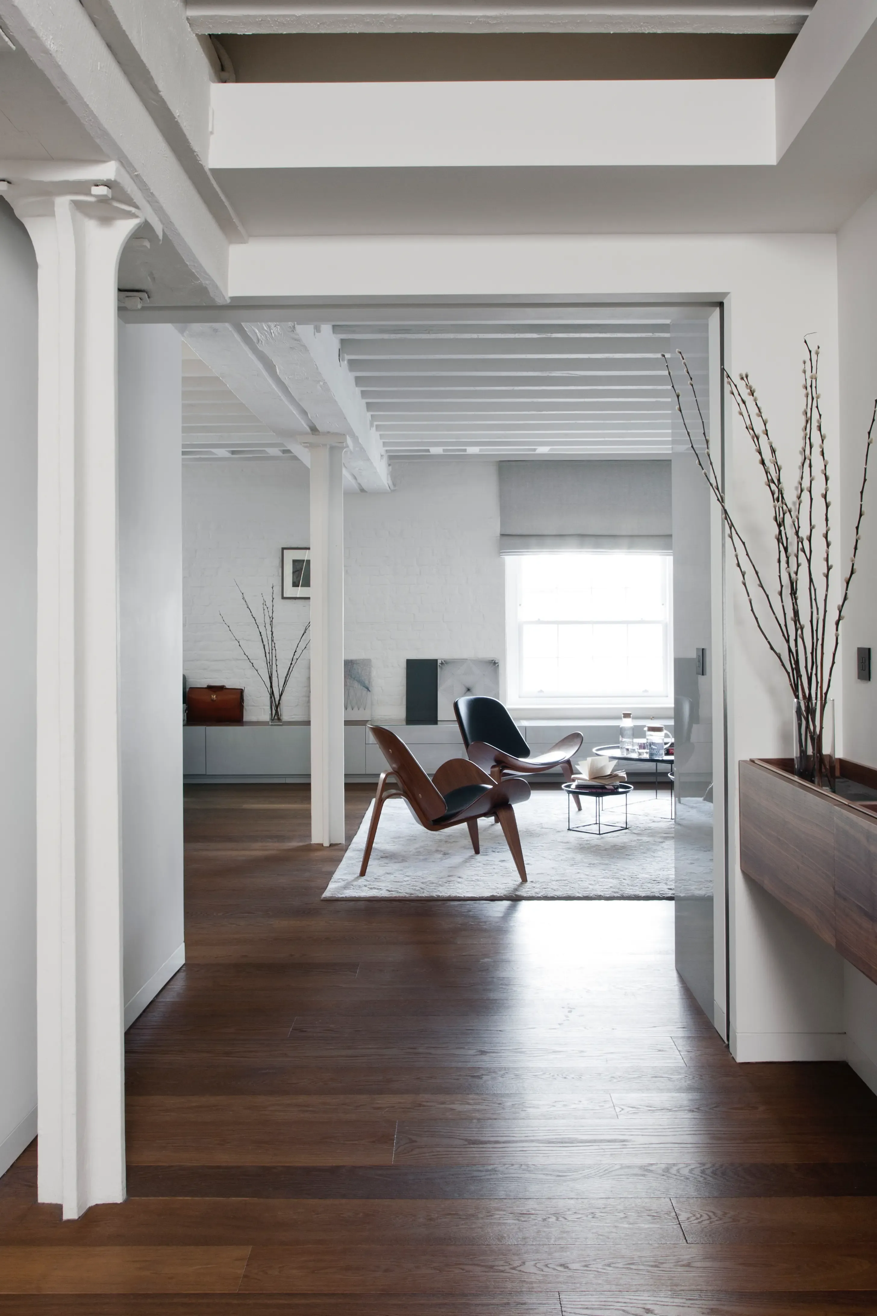 Modern hallway with wooden floors, white beams, and view into bright living area featuring contemporary lounge chairs and minimalist interior design.