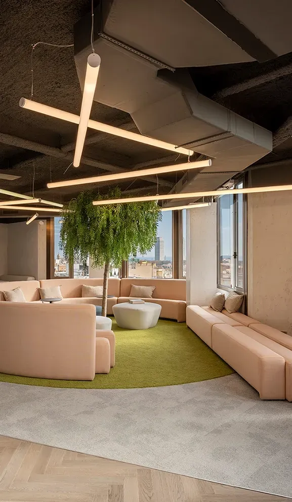 Office lounge with soft lighting, tree centerpiece, and curved sofas offering city views.
