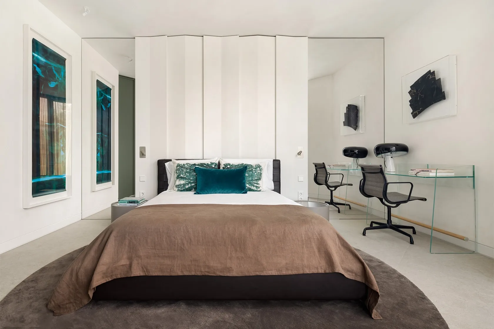 Contemporary bedroom featuring upholstered bed with brown linen bedding, round rug and mirrored wall behind glass desks.