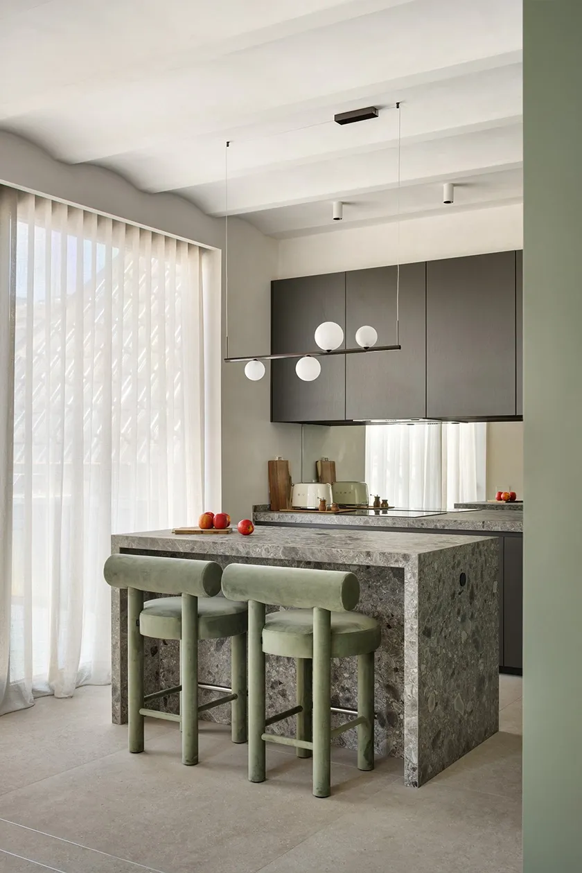 Kitchen island in grey natural stone with sage green velvet bar stools and minimalist pendant lamp with opal glass globes.