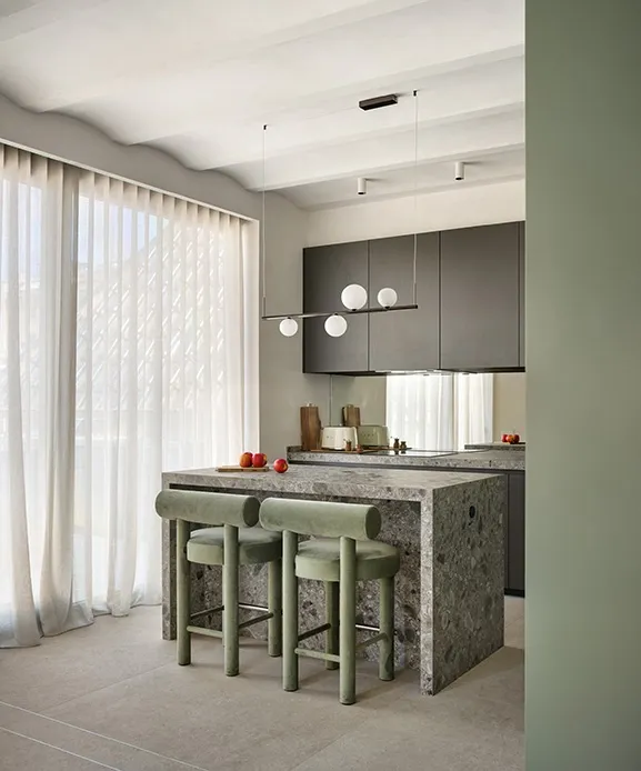 Kitchen island in grey natural stone with sage green velvet bar stools and minimalist pendant lamp with opal glass globes.