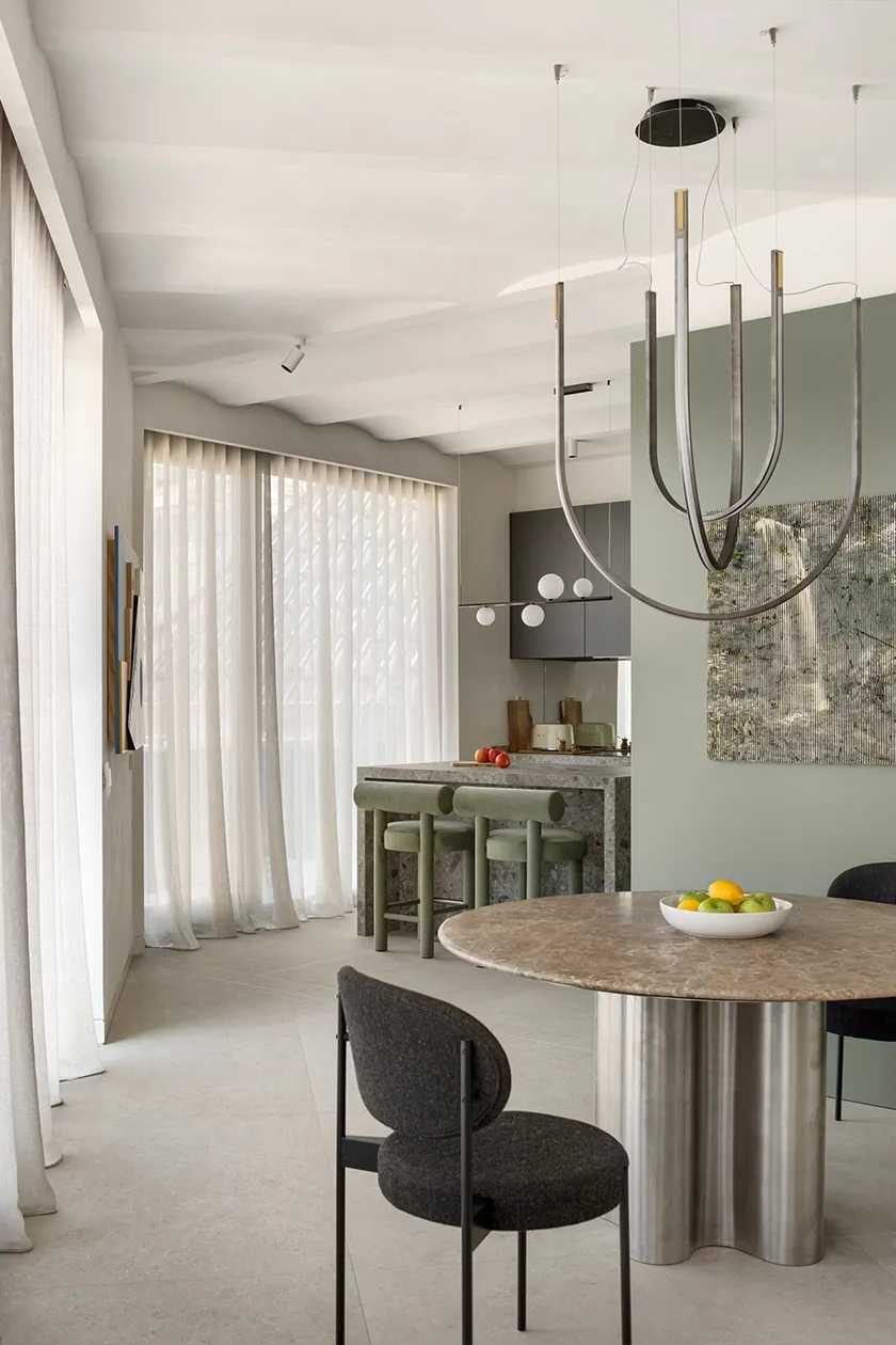 Open-plan dining and living area with round marble table on stainless steel base, dark upholstered chairs and sculptural pendant lighting.
