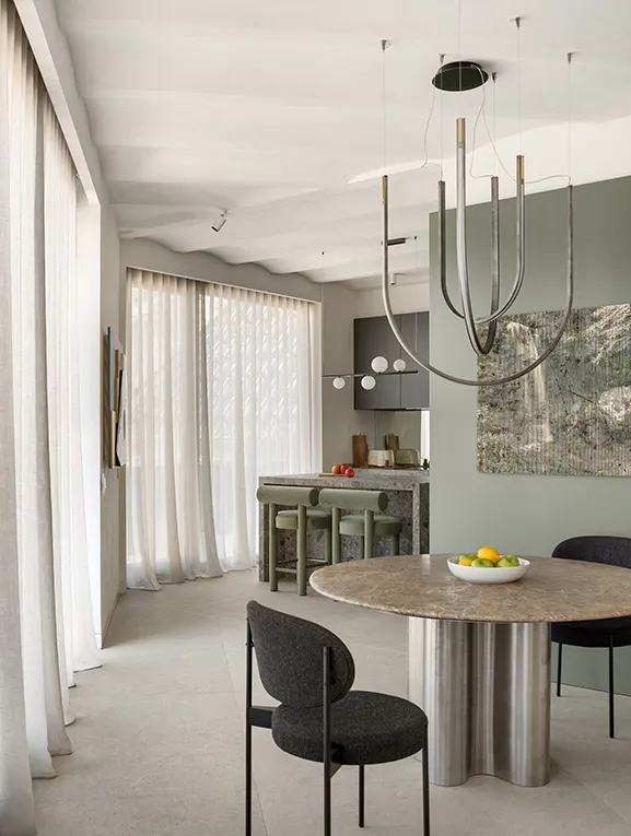 Open-plan dining and living area with round marble table on stainless steel base, dark upholstered chairs and sculptural pendant lighting.