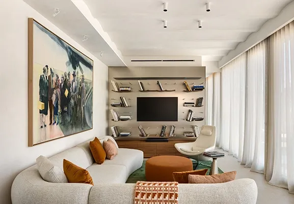Curved sectional sofa with warm-toned cushions facing large artwork and integrated metal bookshelf with TV.