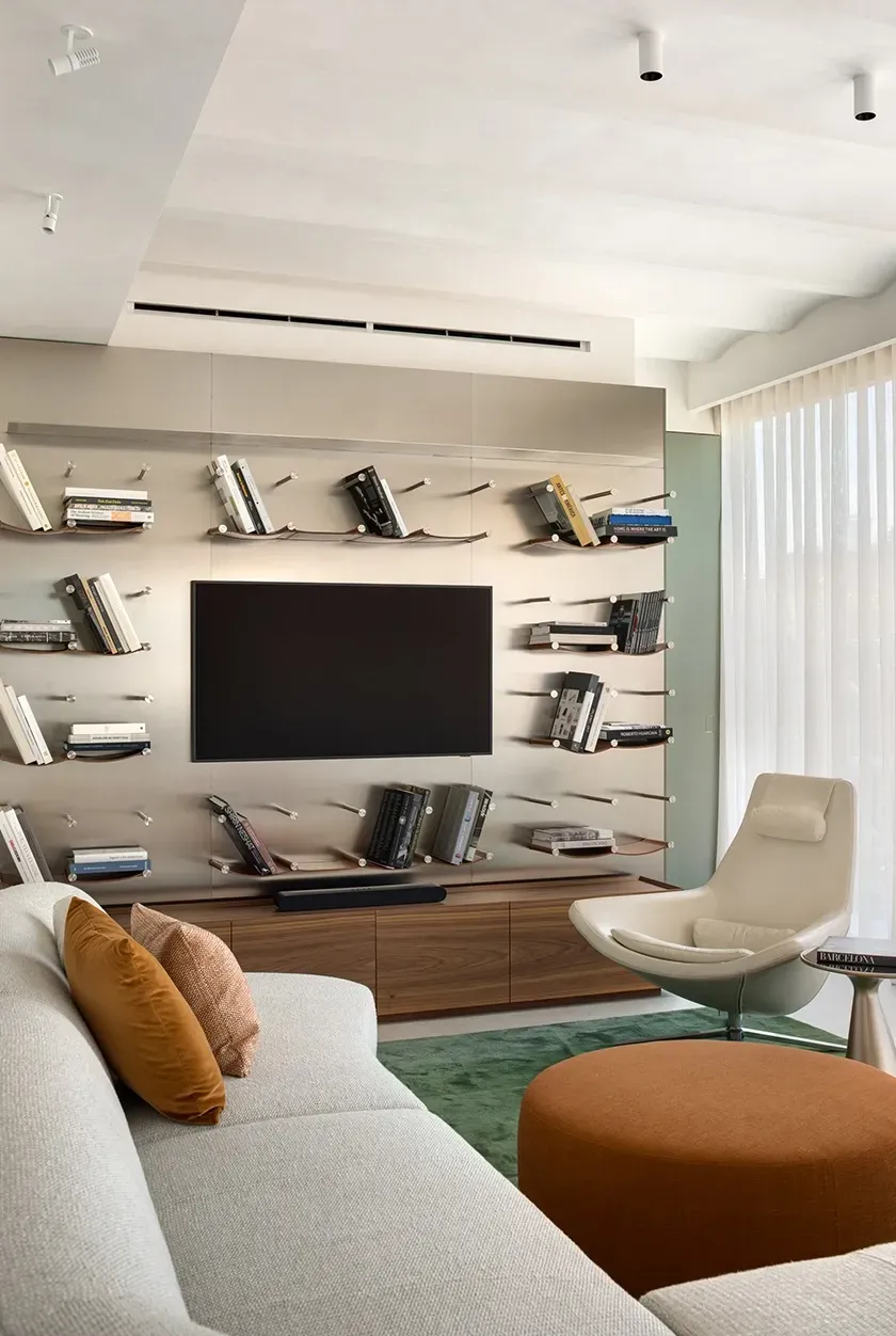 Living room with curved upholstered sofa, green area rug, orange ottoman and wall-mounted metal bookshelf system around TV.