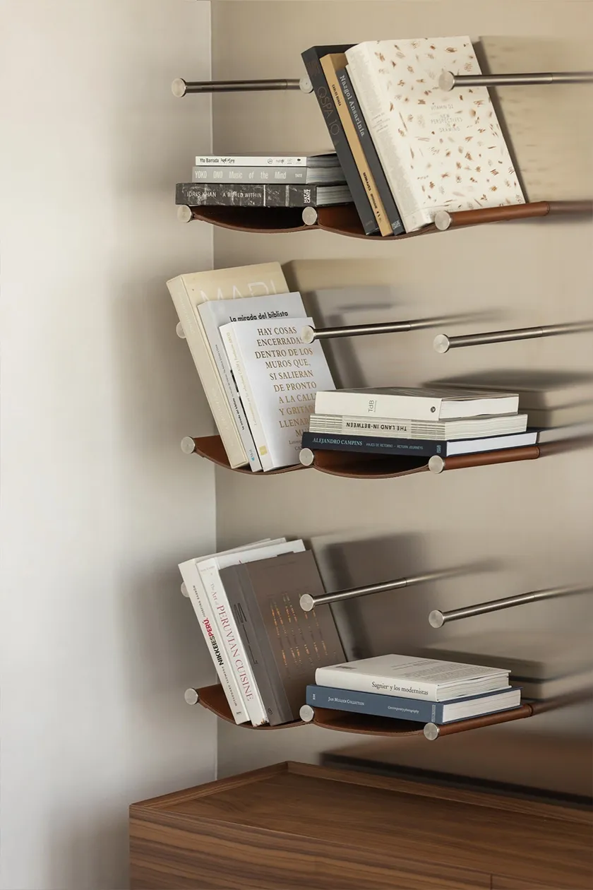 Floating leather and metal bookshelves with assorted design books mounted on light wall.