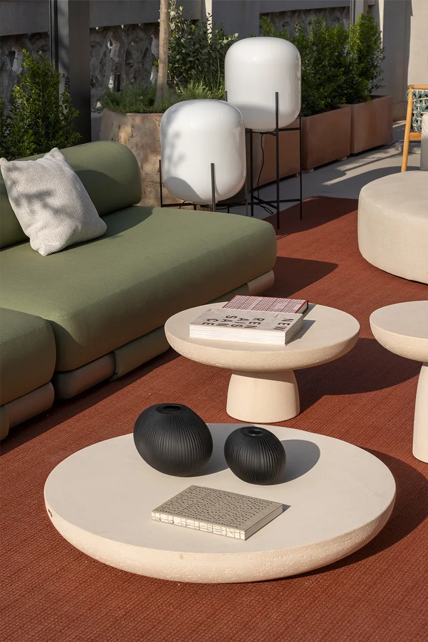 Outdoor lounge area with green upholstered modular sofa, circular stone coffee tables and matte black decorative vases on terracotta rug.