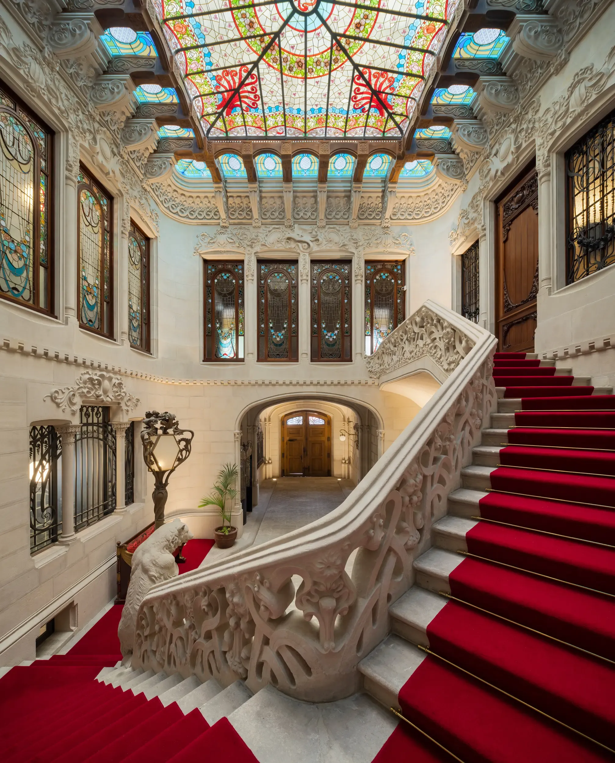 Red-carpeted staircase with intricate stone balustrade and stained glass ceiling in elegant architectural setting.