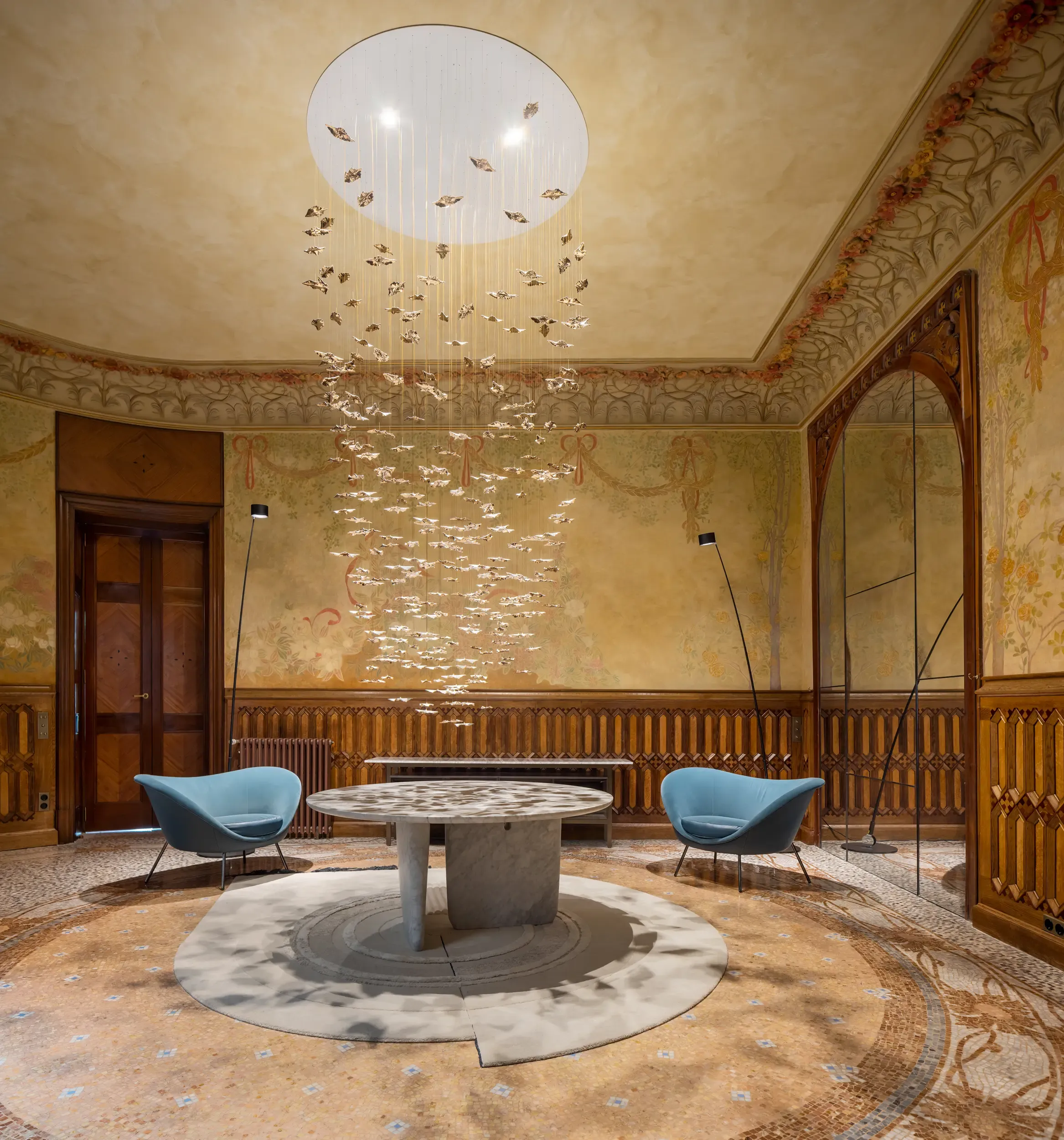 Artistic lounge with frescoed walls, marble table, modern chairs, and cascading sculptural light installation.