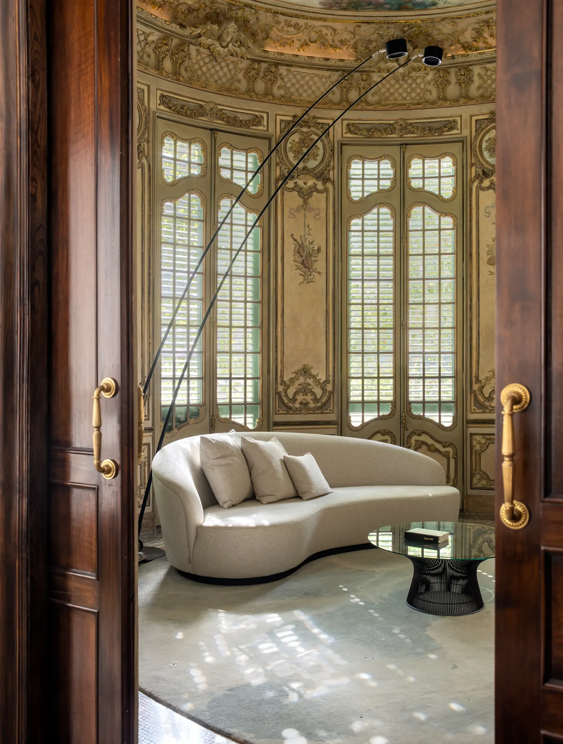 Intimate view of curved sofa and glass table under frescoed ceiling with tall windows.