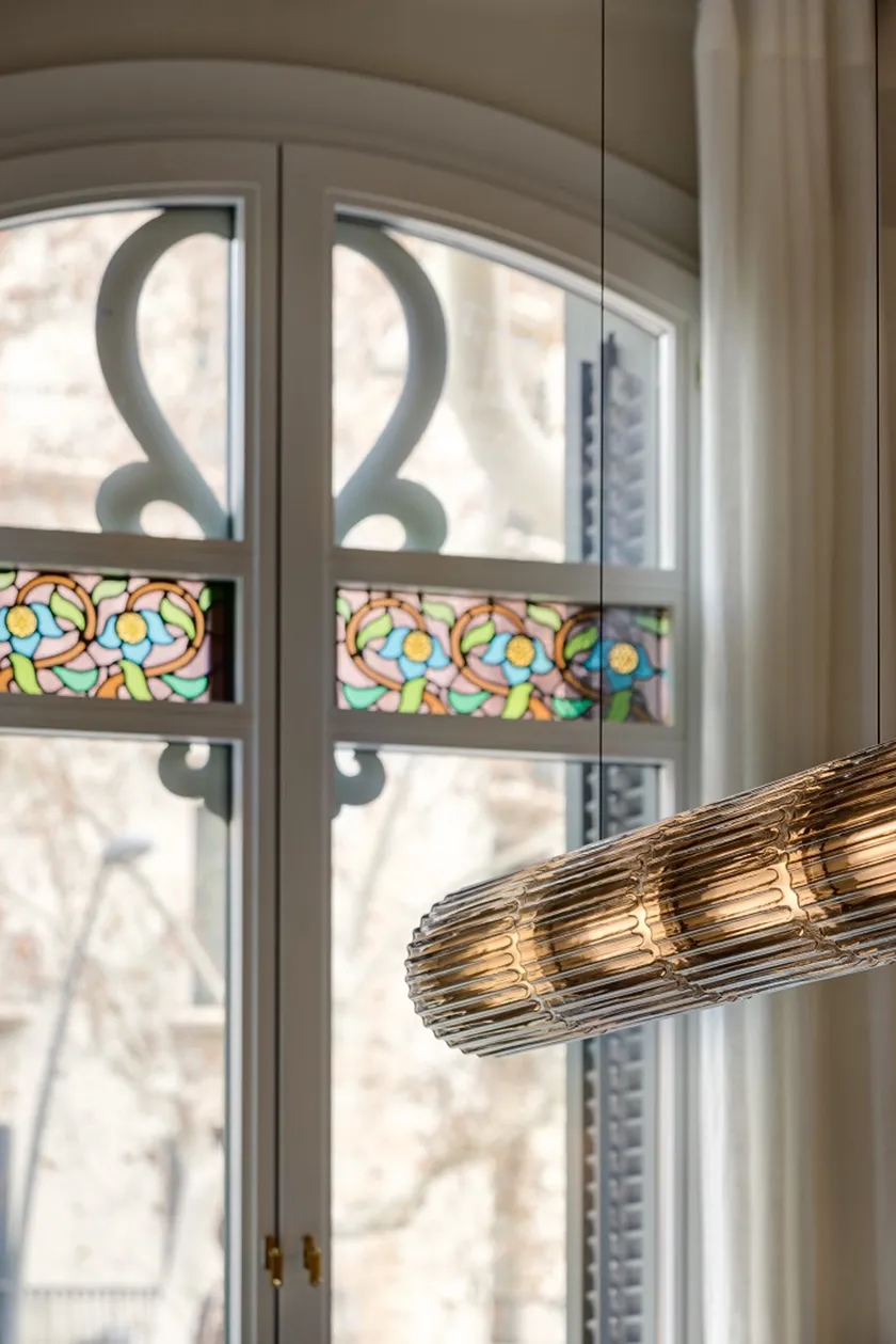 Detalle de una lámpara colgante contemporánea junto a una ventana con vidrieras florales modernistas en Casa Oller.