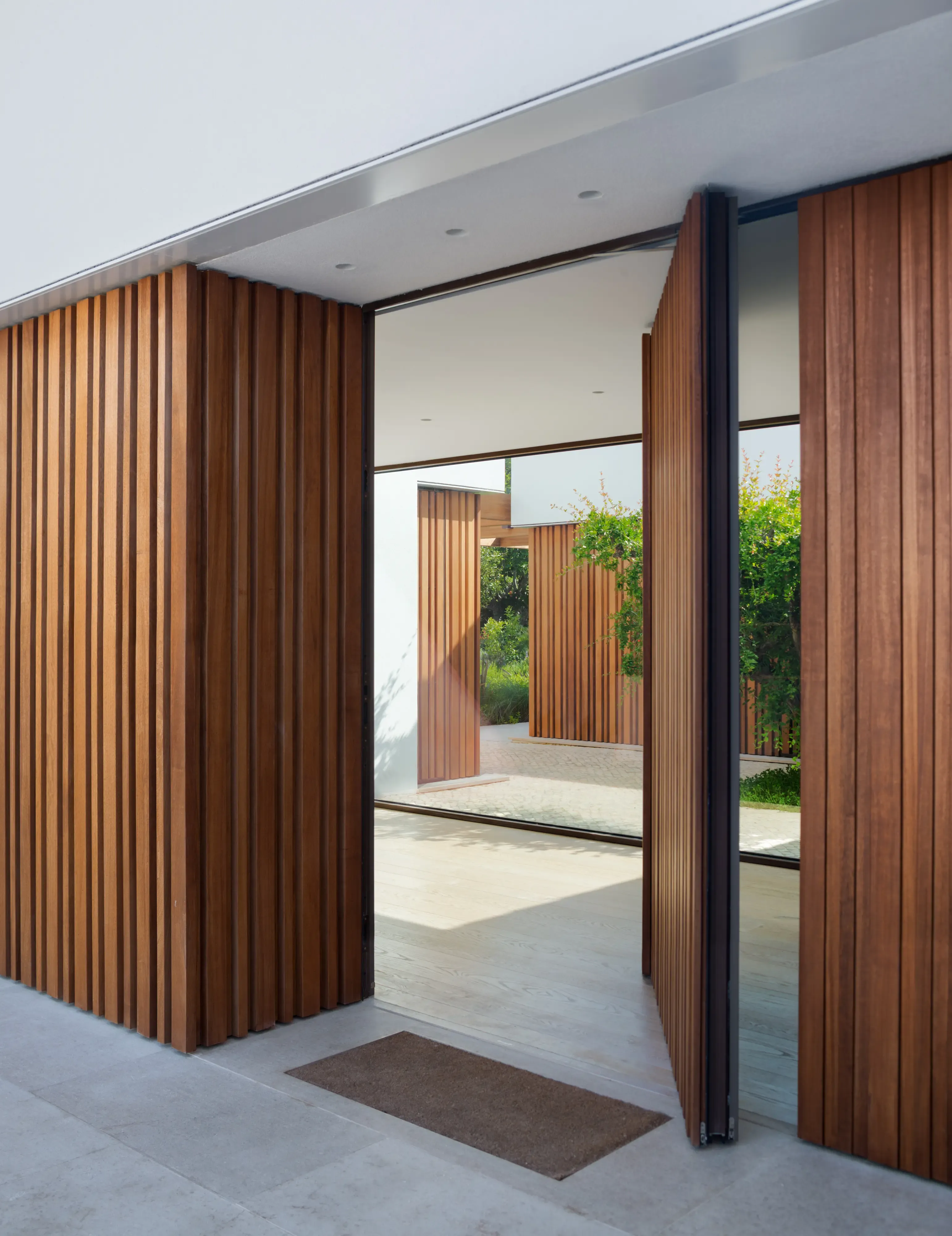 Pivoting wooden door opening to reveal a contemporary entrance framed by vertical slats and lush landscaped garden beyond.