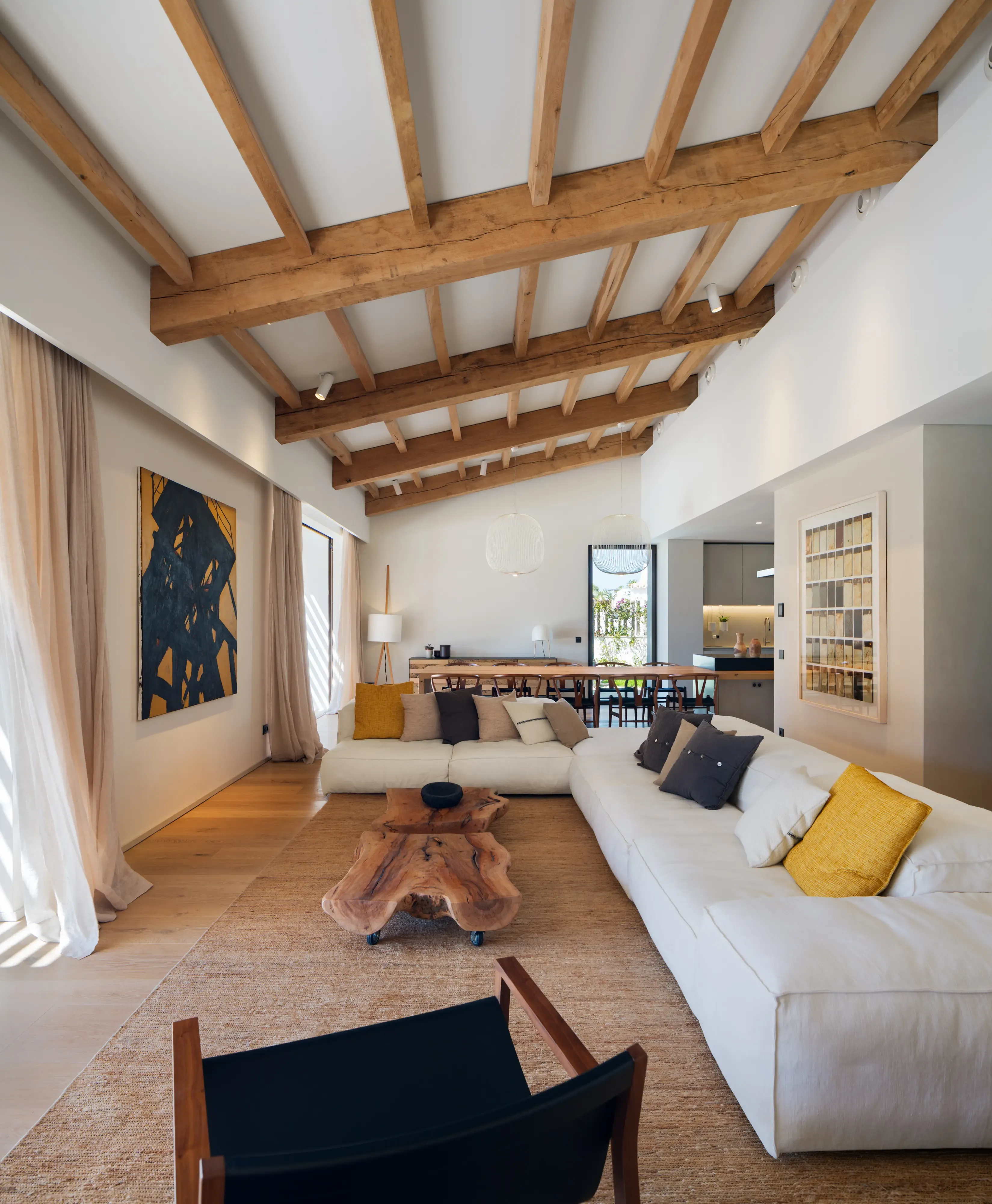 Spacious living room with exposed wooden beams, large white sofas, rustic wood coffee table, and modern art accentuating warm natural tones.