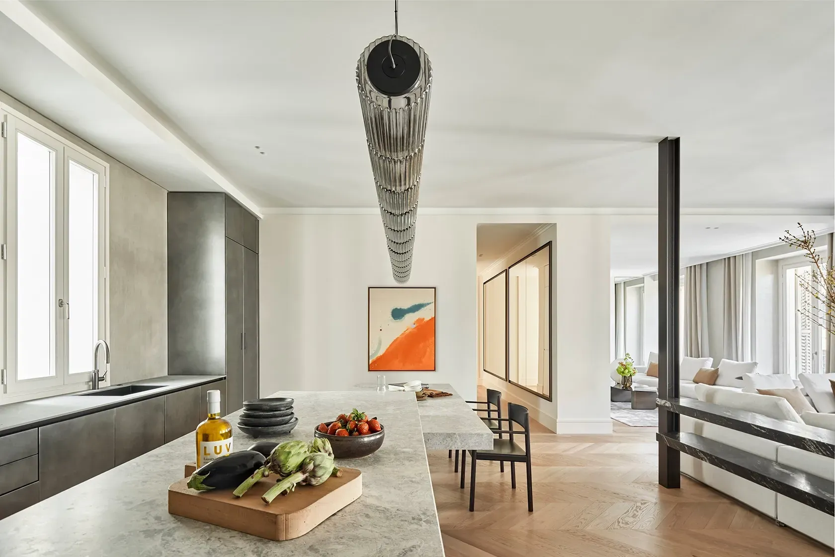 Open kitchen space with textured grey island, organic still life arrangement, and connection to a bright living area with neutral-toned furniture.