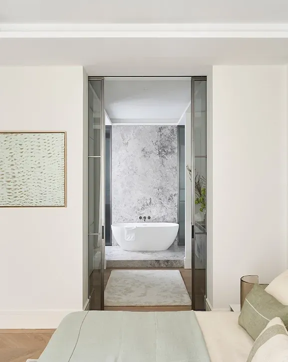 Master bedroom with upholstered wall paneling and neutral tones, leading to a marble-finished ensuite bathroom with freestanding tub and soft lighting.