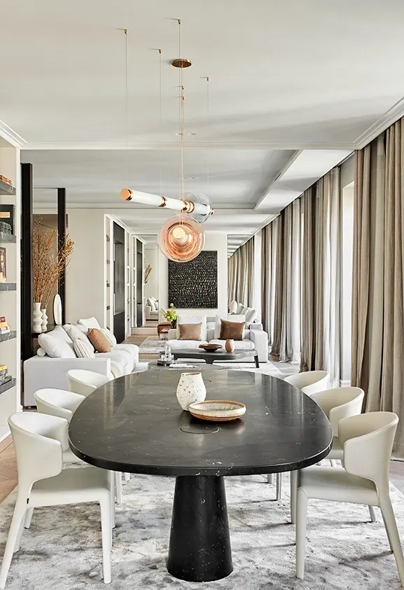 Open-plan dining and living room with statement chandelier, large windows, elegant curtains, and sophisticated art pieces in a luminous, serene interior.