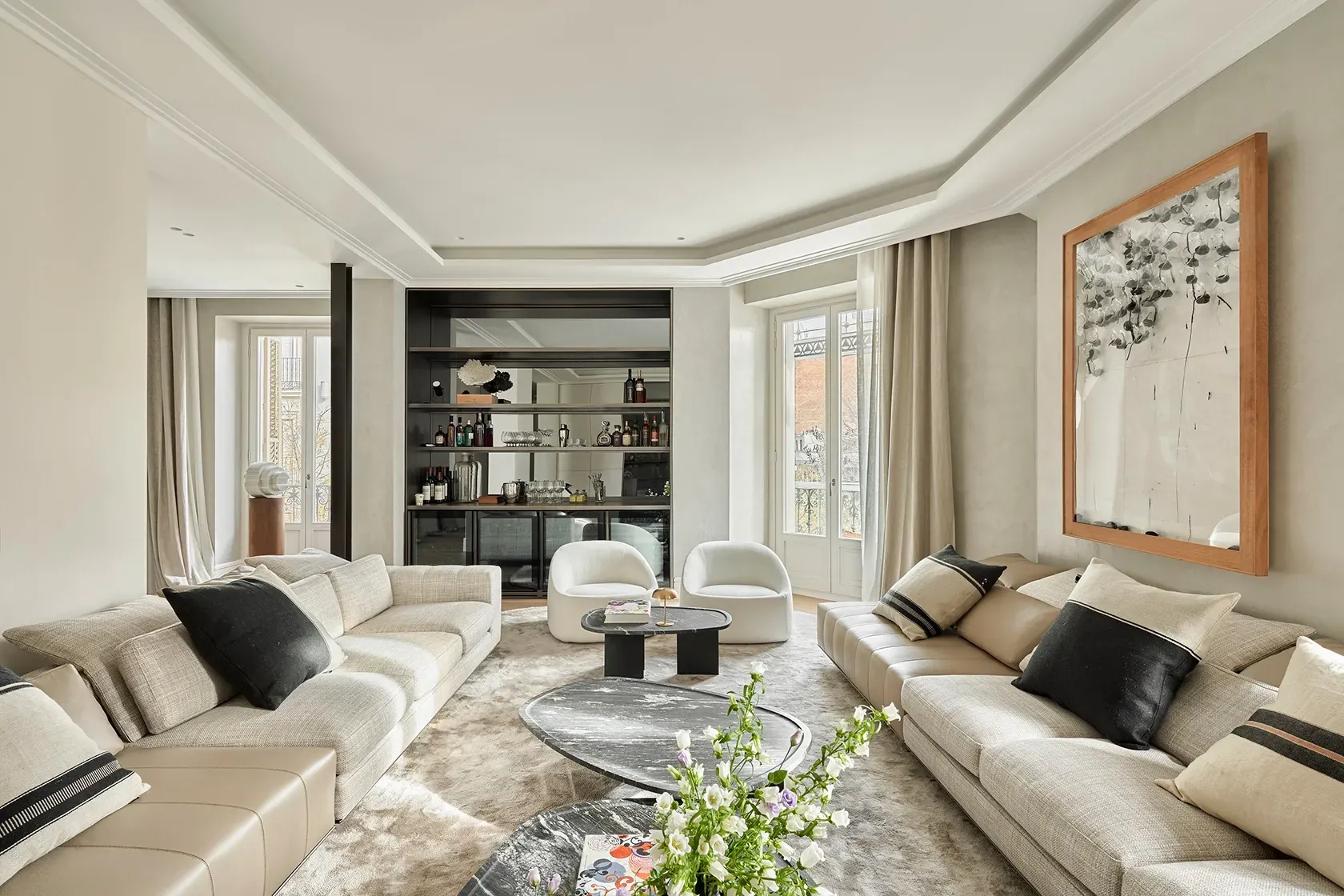 Luxury living room with light neutral sofas, custom bar, modern artwork, and large windows filling the space with warm, natural daylight.
