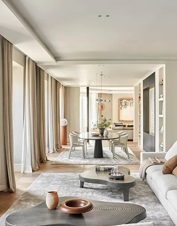Living area with pale sofas, sculptural decor, and a view towards the dining space, framed by elegant drapes and soft ambient light throughout.