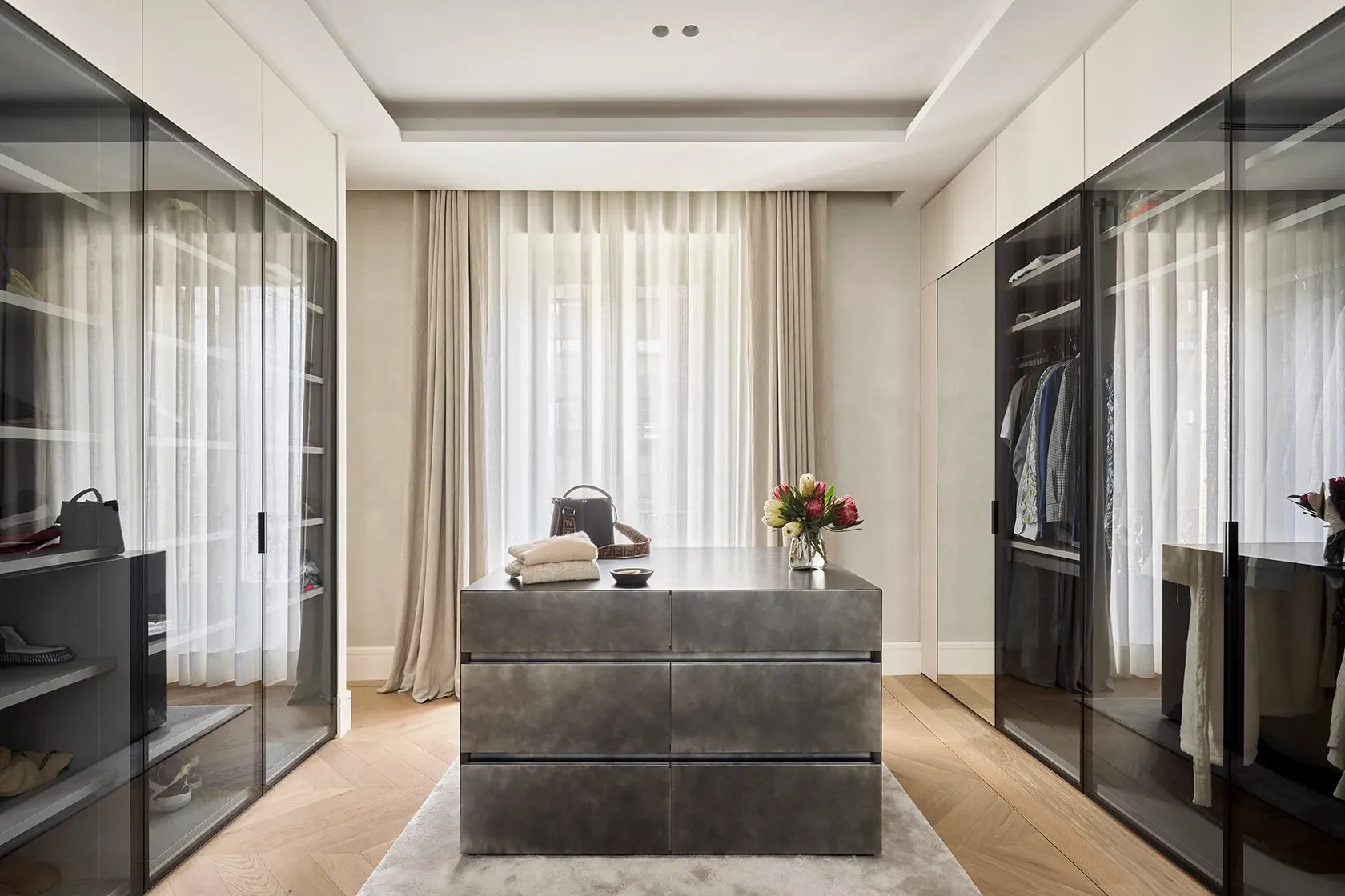 High-end walk-in closet with glass-front wardrobes, metallic island dresser, and natural light streaming through sheer curtains and neutral textiles.