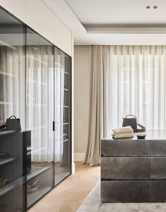 High-end walk-in closet with glass-front wardrobes, metallic island dresser, and natural light streaming through sheer curtains and neutral textiles.