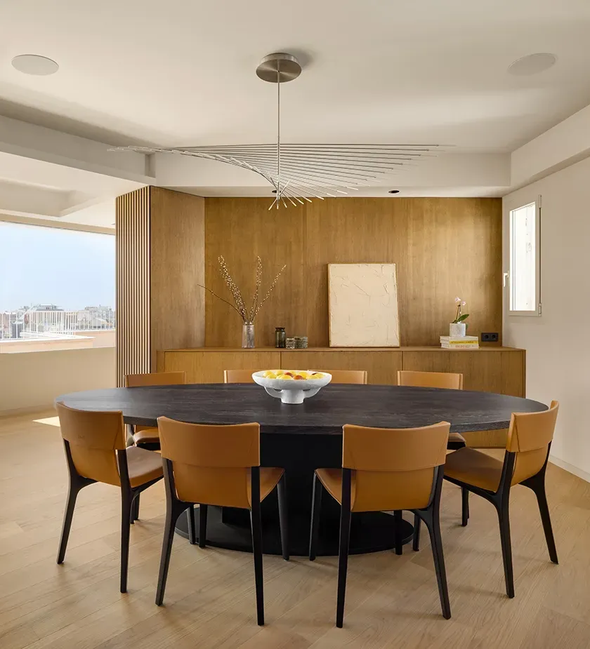 Elegant dining room with dark oval table, tan leather chairs, and sculptural ceiling light, set against wood paneling and a large window view.