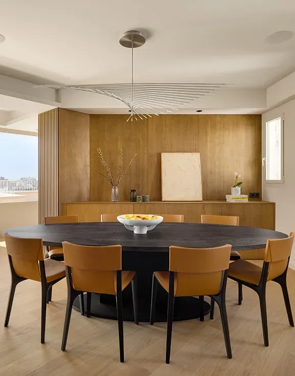Elegant dining room with dark oval table, tan leather chairs, and sculptural ceiling light, set against wood paneling and a large window view.