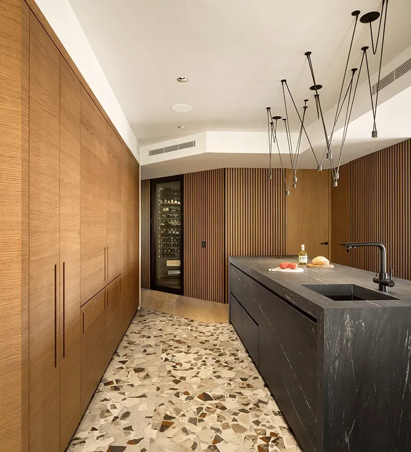 Sleek kitchen with terrazzo flooring, fluted wood walls, and black marble island, featuring integrated wine storage and modern lighting.