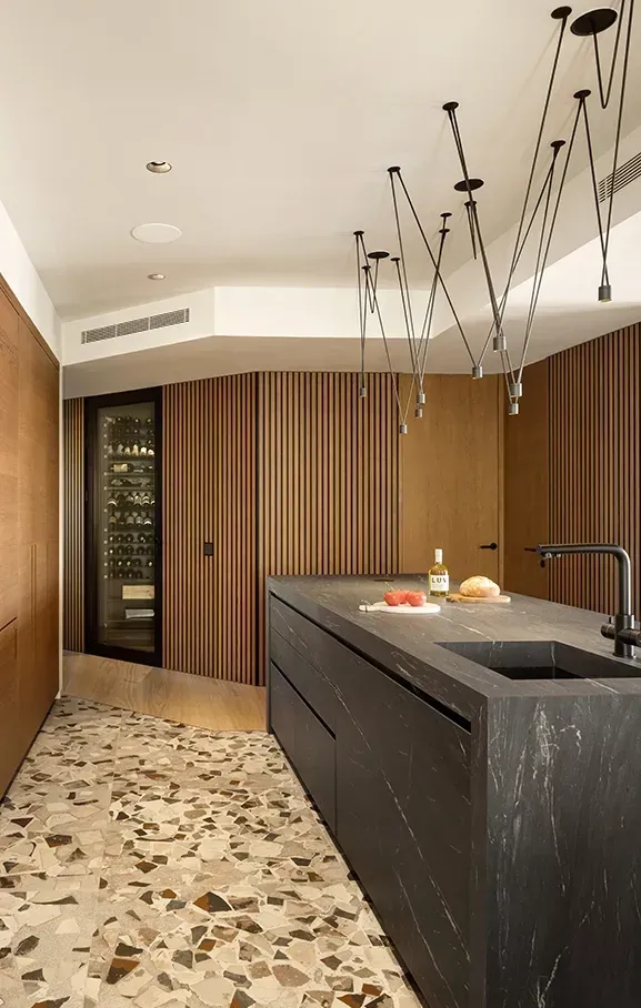 Sleek kitchen with terrazzo flooring, fluted wood walls, and black marble island, featuring integrated wine storage and modern lighting.