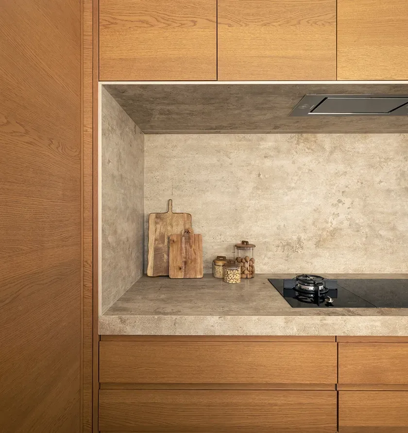 Neutral-toned kitchen nook with textured stone backsplash, wood cabinetry, and open shelves holding cutting boards, jars, and spices.
