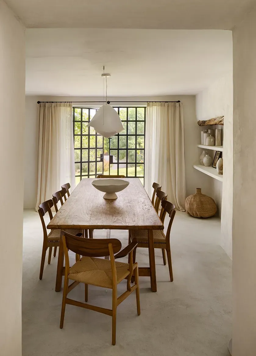 Warm dining room with rustic wooden table, woven chairs, ceramic decor, and soft light flowing through black-grid windows and linen curtains.