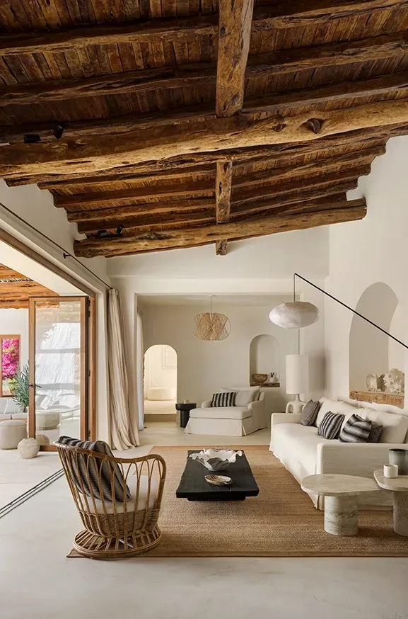 Open-plan living area with exposed wooden beams, neutral furnishings, and a seamless transition to the outdoor lounge with natural light.
