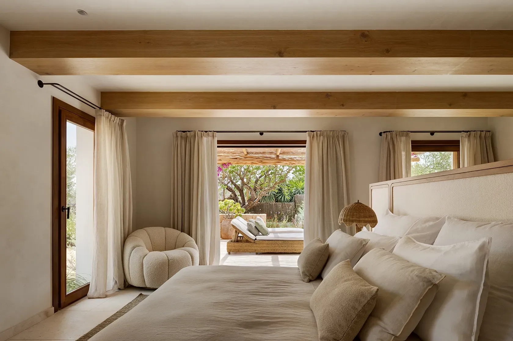 Master bedroom with light wooden beams, soft textures, and direct access to a lush terrace with outdoor lounge and blooming bougainvillea.