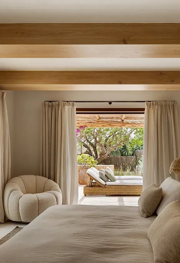 Master bedroom with light wooden beams, soft textures, and direct access to a lush terrace with outdoor lounge and blooming bougainvillea.