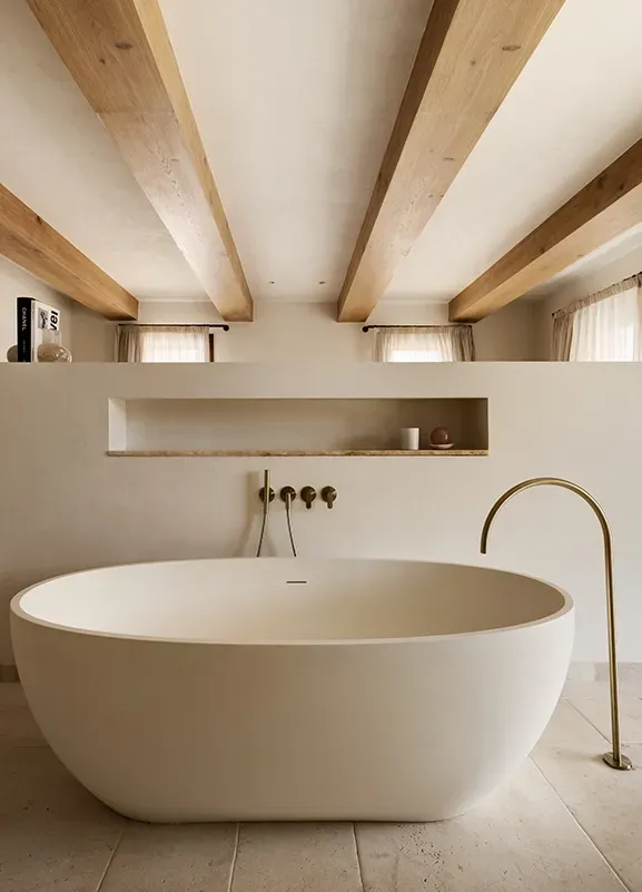 Refined bathroom with sculptural freestanding tub, light wooden ceiling beams, and warm natural light for a tranquil wellness experience.