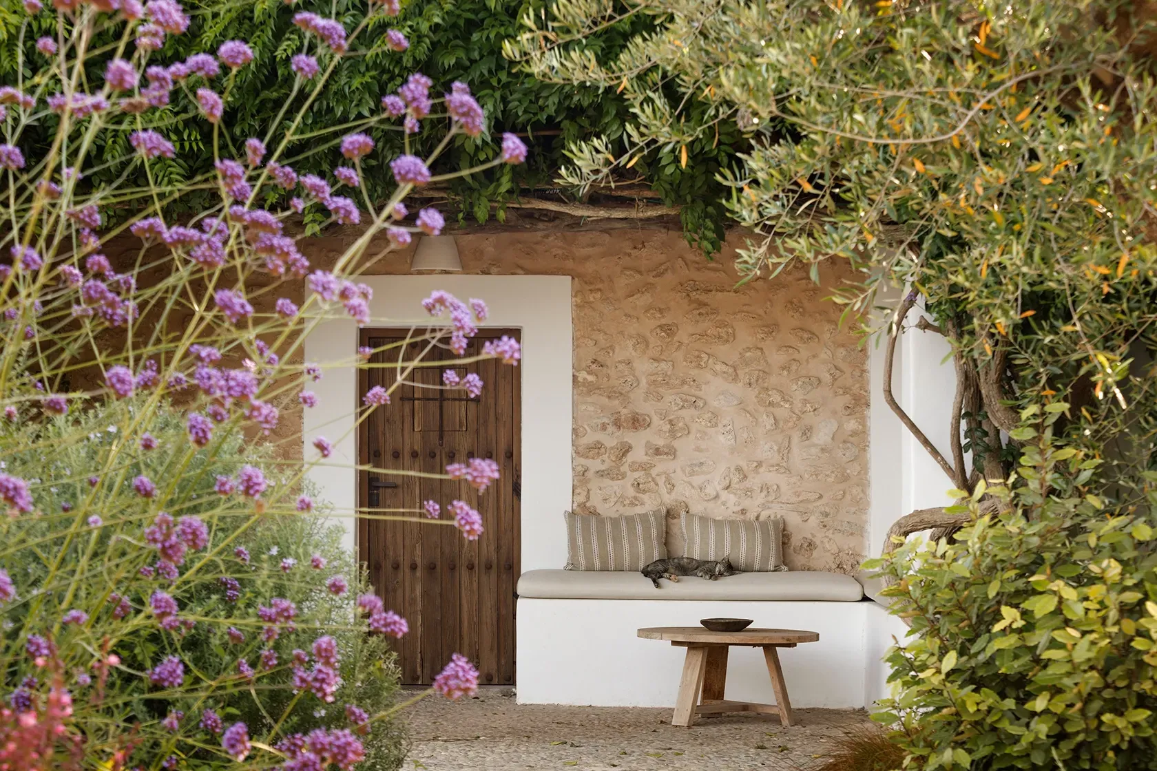 Charming garden nook with stone walls, wooden door, bench seating, and blooming purple flowers—perfect for peaceful moments outdoors.
