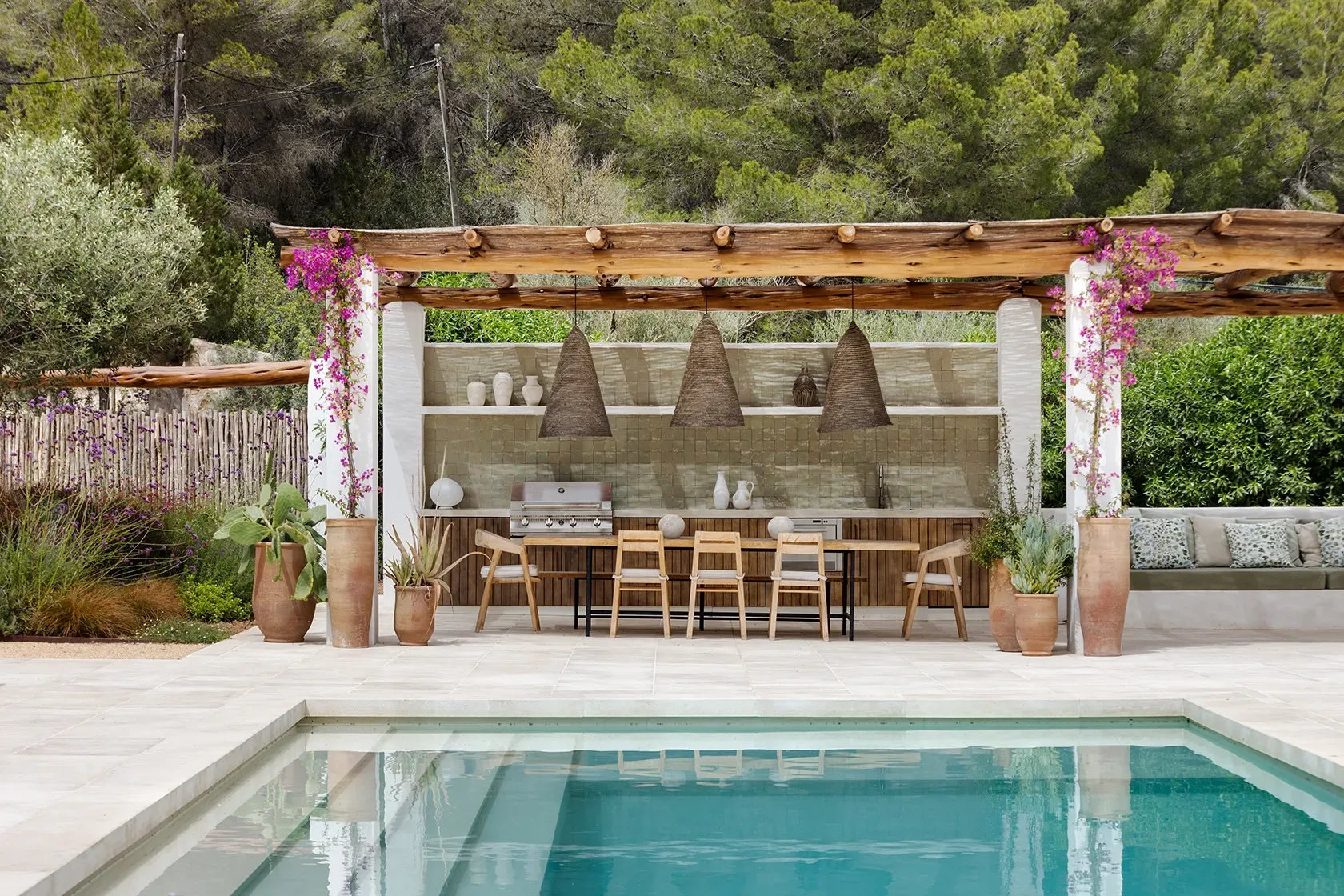 Poolside lounge with natural stone flooring, pergola-covered dining, and lush Mediterranean landscaping for a luxury outdoor experience.