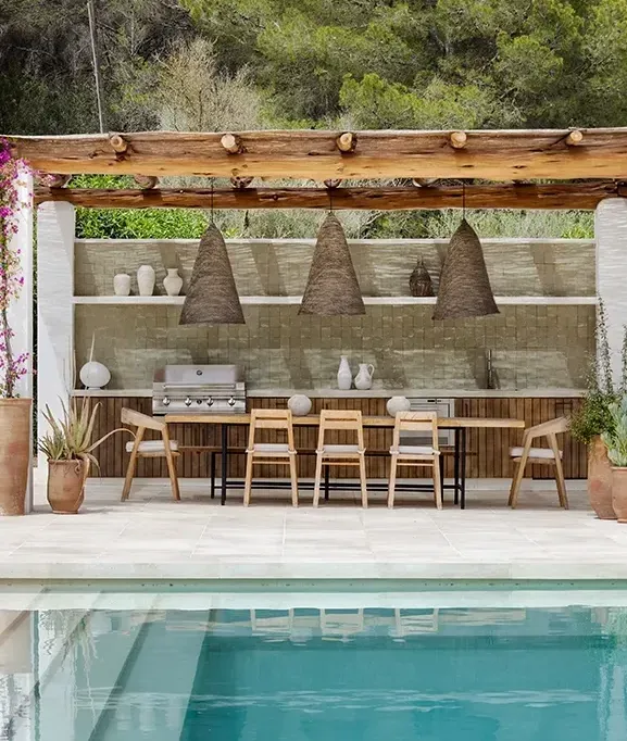 Poolside lounge with natural stone flooring, pergola-covered dining, and lush Mediterranean landscaping for a luxury outdoor experience.