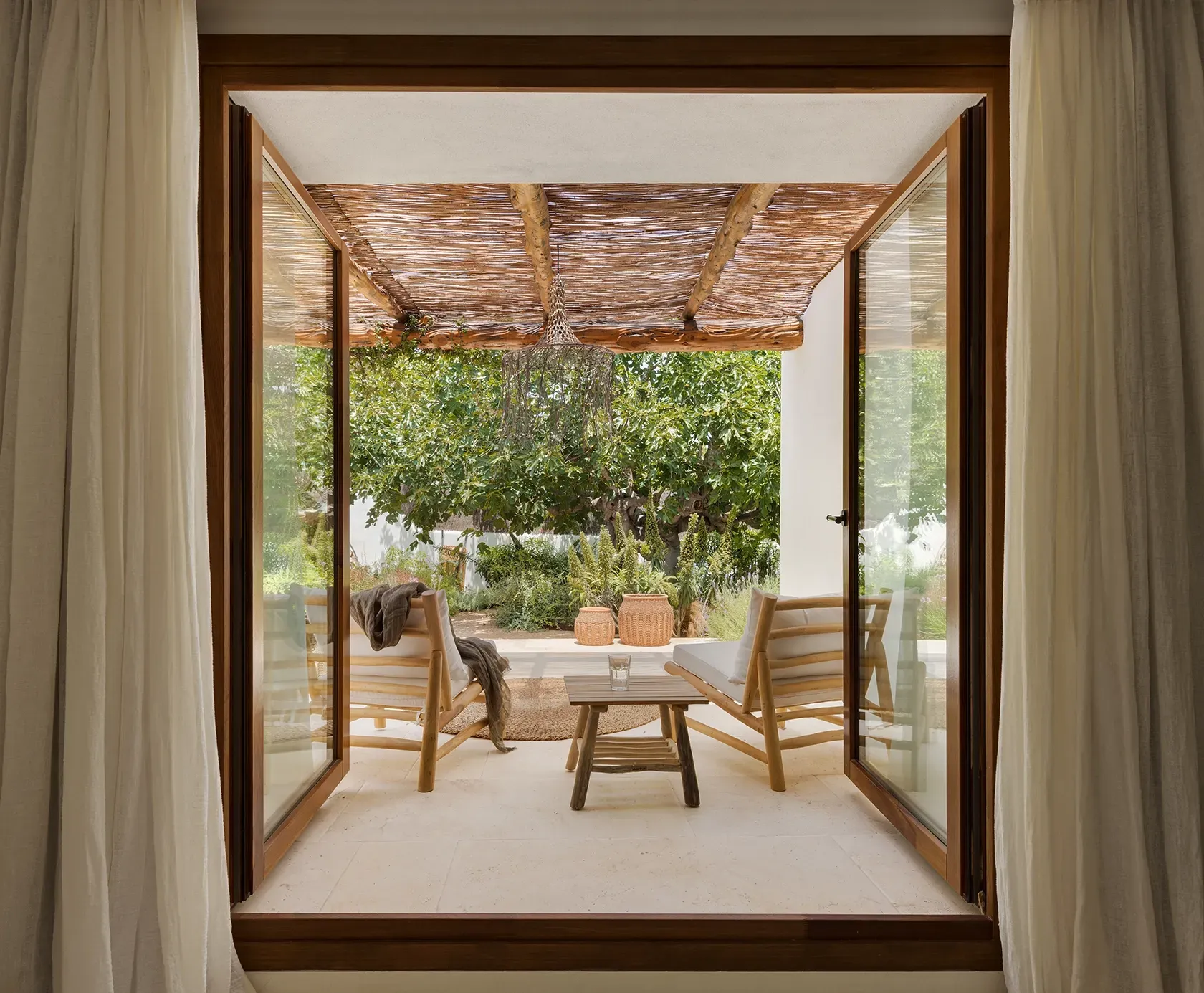 Wood-framed glass doors open to a private terrace with a natural pergola, cozy seating, and lush greenery—blending indoor and outdoor living.