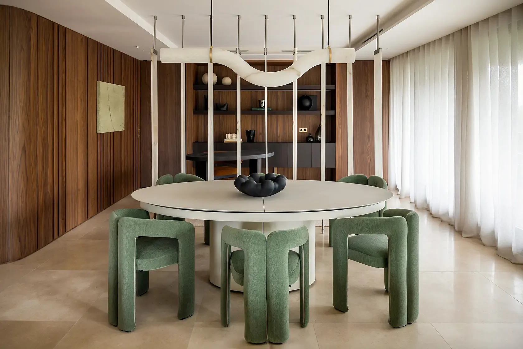 Contemporary dining room with a round statement table, upholstered green chairs, sculptural suspended lighting and translucent stone partitions, framed by custom walnut paneling and natural light.