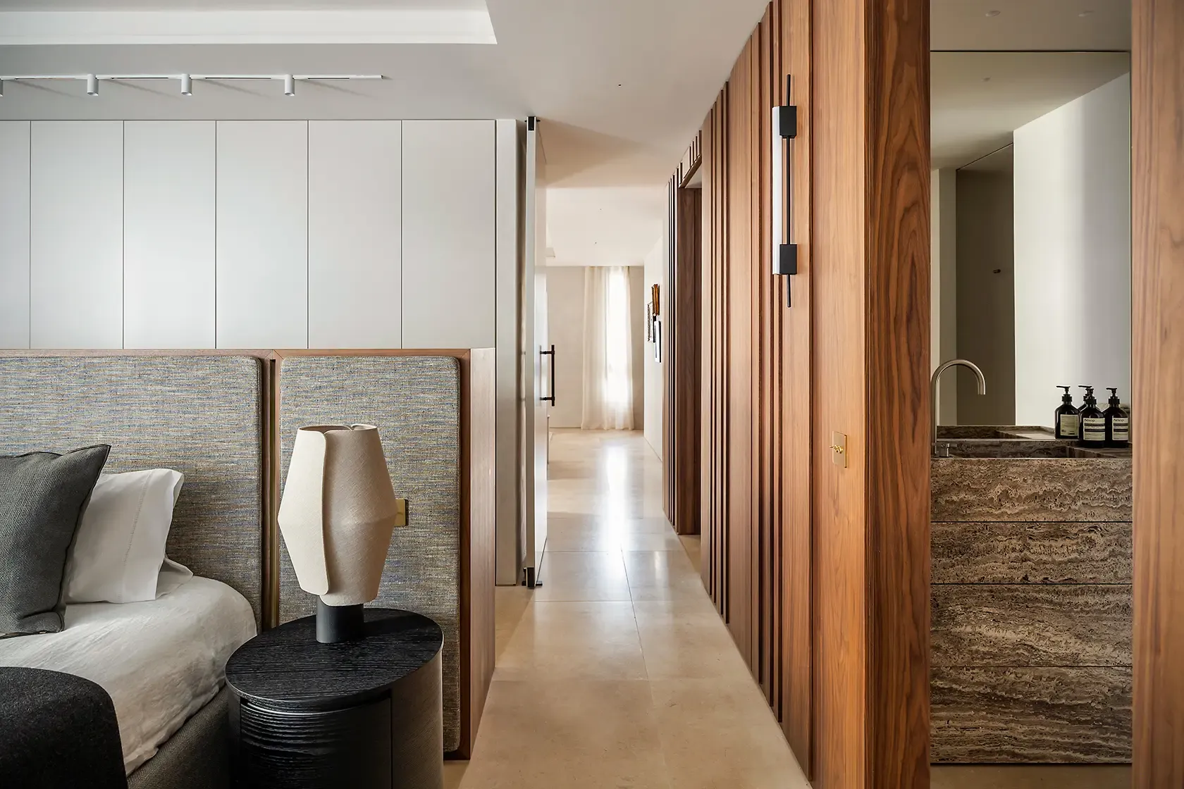 Guest bedroom with an upholstered headboard, custom walnut wall paneling, integrated bathroom vanity in natural stone and a warm contemporary material palette.