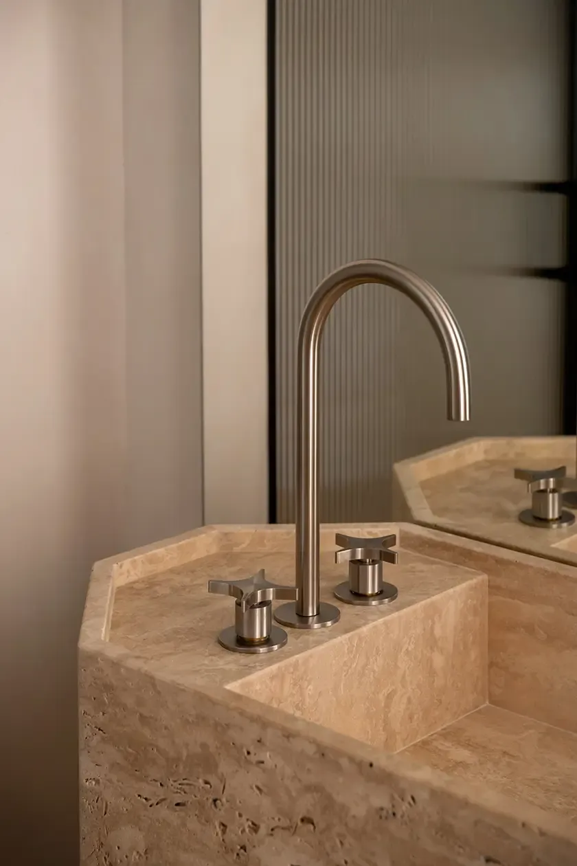 Close-up of a custom travertine vanity with integrated sink, brushed metal faucet and textured glass backdrop, showcasing refined craftsmanship and natural stone detailing.