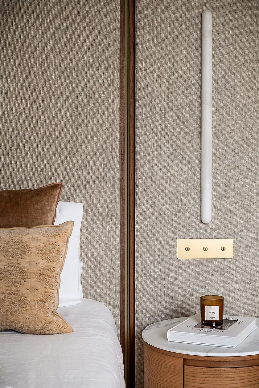Master bedroom detail featuring a textured upholstered headboard, marble-topped bedside table, warm wood finishes and integrated wall lighting, creating a calm and refined atmosphere.