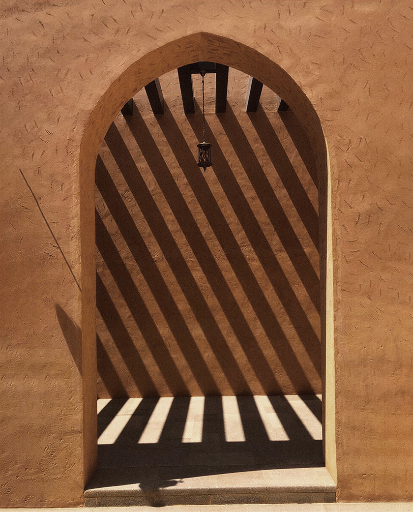 Textured earthen archway with dramatic striped shadows cast by a pergola, highlighting the interplay of light, geometry and desert-inspired architecture.