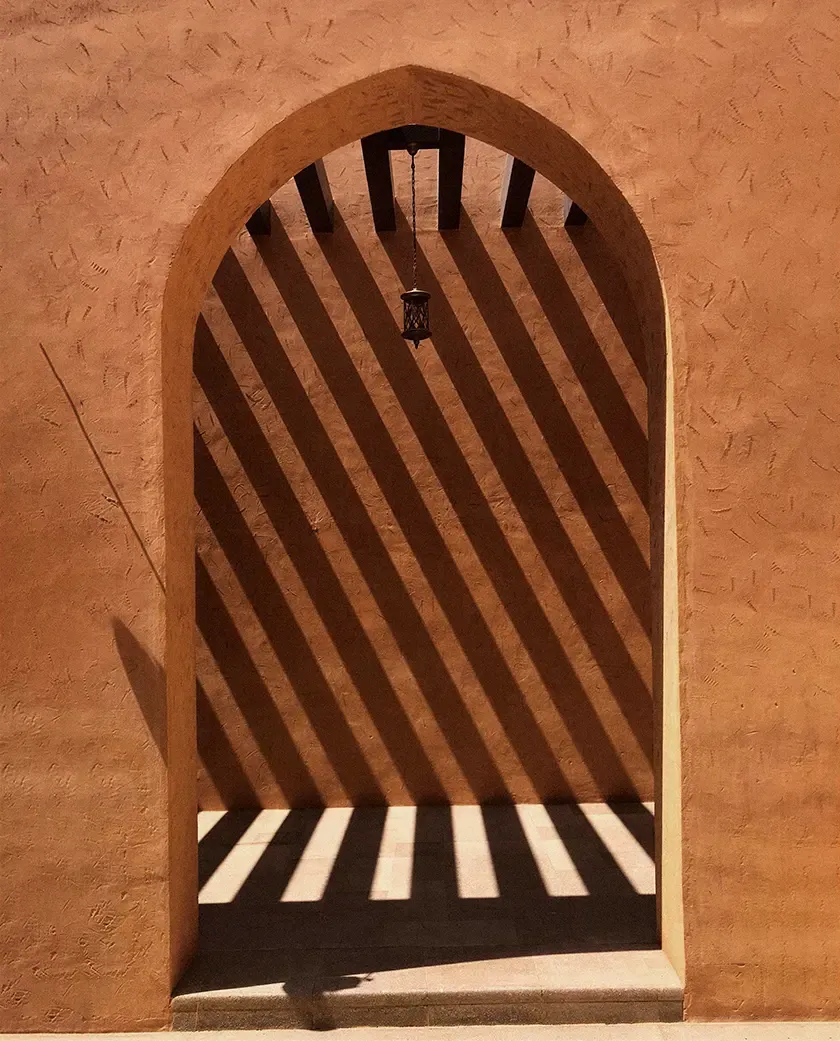Textured earthen archway with dramatic striped shadows cast by a pergola, highlighting the interplay of light, geometry and desert-inspired architecture.