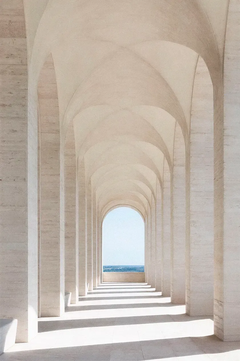 Monumental vaulted colonnade in light stone, featuring rhythmic arches, soft natural light and a framed view of the sea beyond.