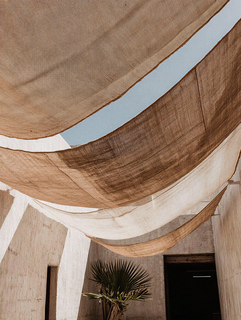 Layered fabric shade sails suspended above a desert-inspired courtyard, filtering natural light across textured plaster walls and tropical planting.