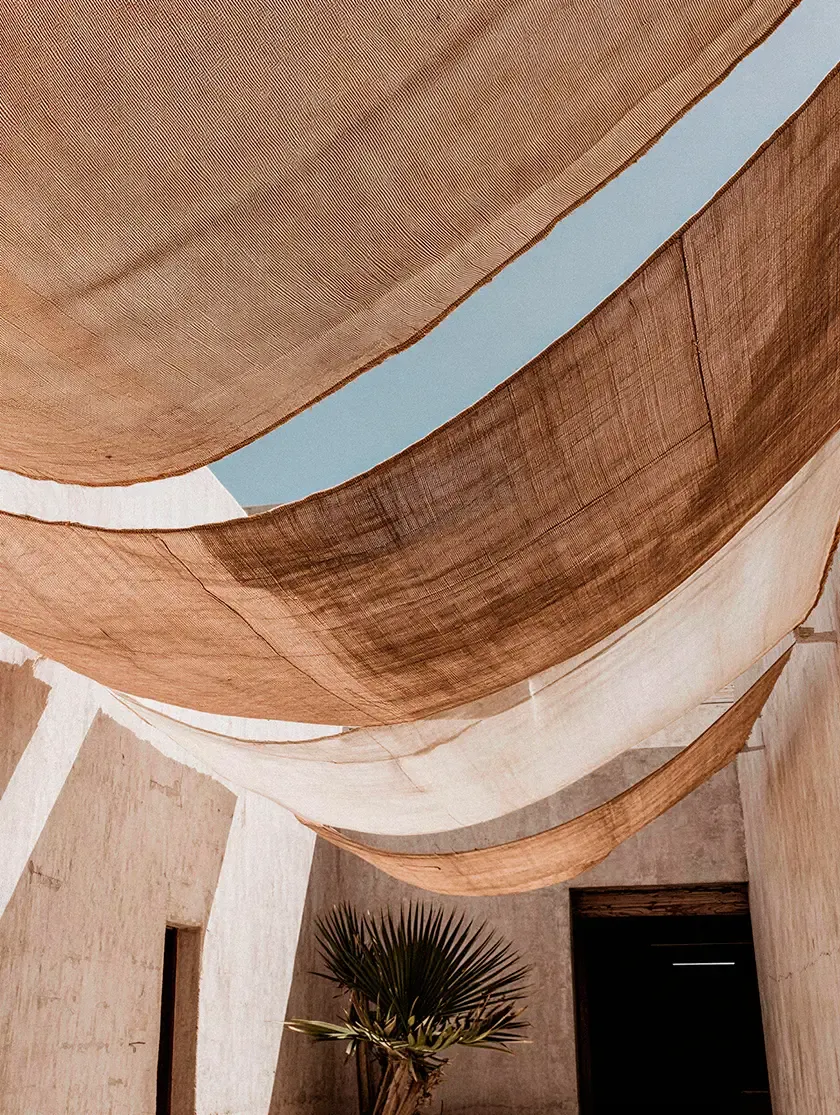 Layered fabric shade sails suspended above a desert-inspired courtyard, filtering natural light across textured plaster walls and tropical planting.