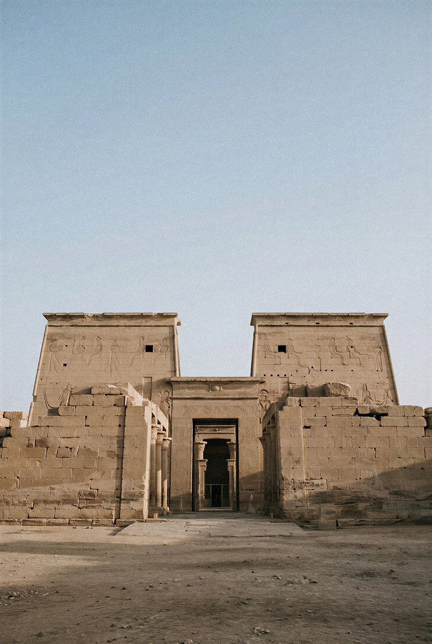 Ancient Egyptian stone temple facade with monumental entrance, carved reliefs and weathered masonry set against a clear desert sky.