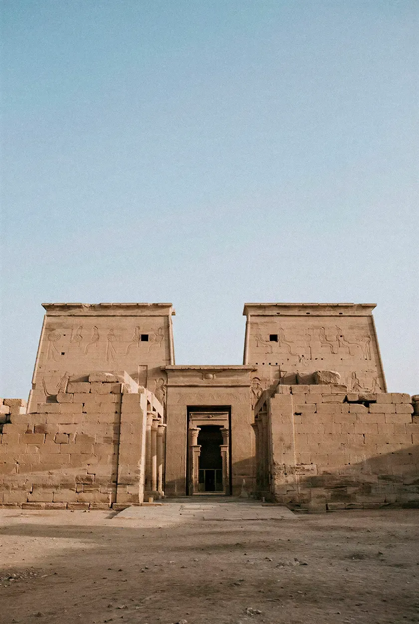 Ancient Egyptian stone temple facade with monumental entrance, carved reliefs and weathered masonry set against a clear desert sky.