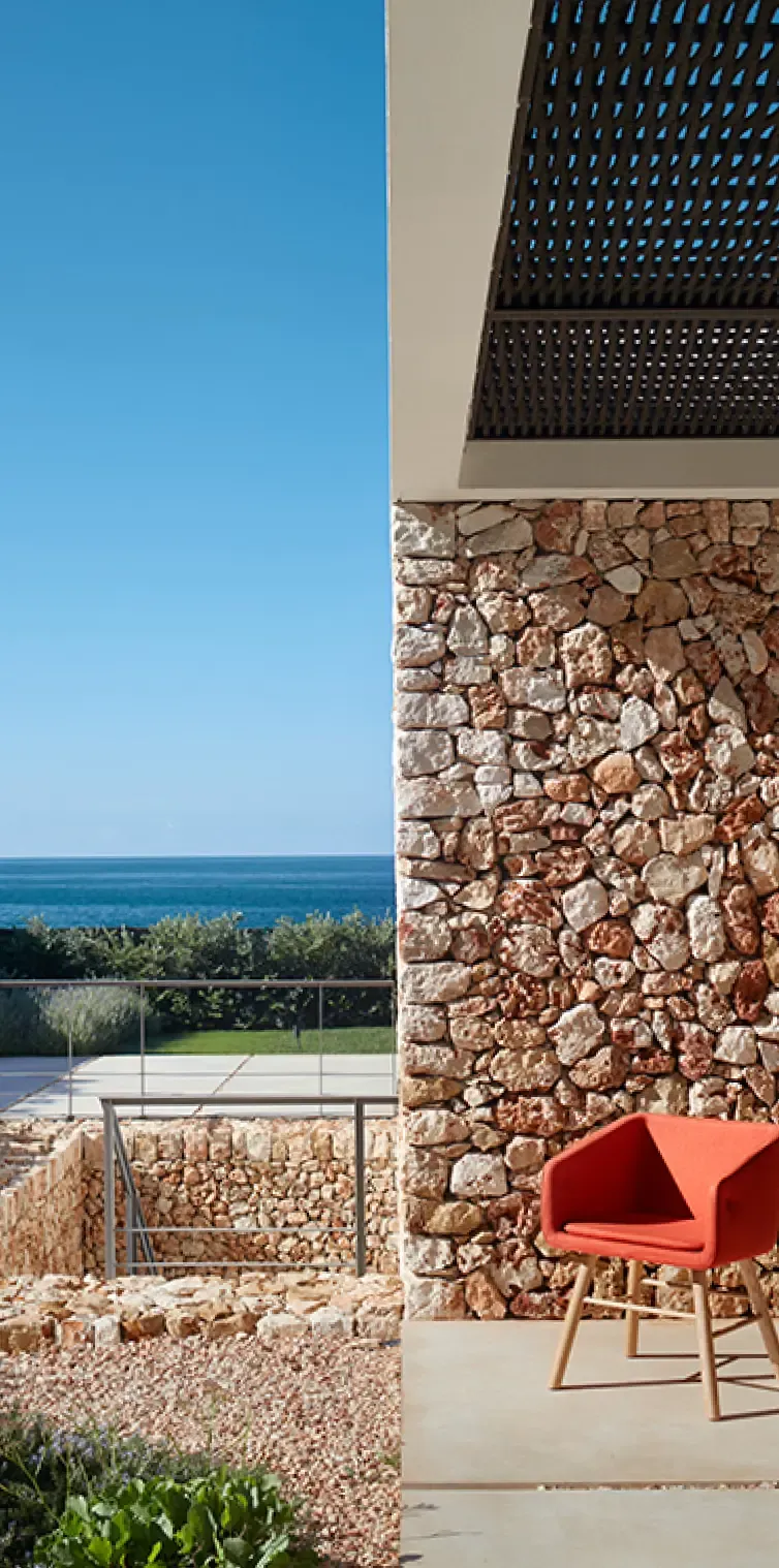 Exterior view of a Mediterranean stone wall with a red chair and ocean backdrop, blending coastal and contemporary design.